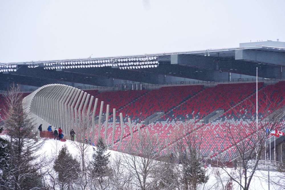 A photo showing the hill at Lansdowne Park in the winter with families tobogganing. The art piece “Moving Surfaces” is shown with the north stands filling the background