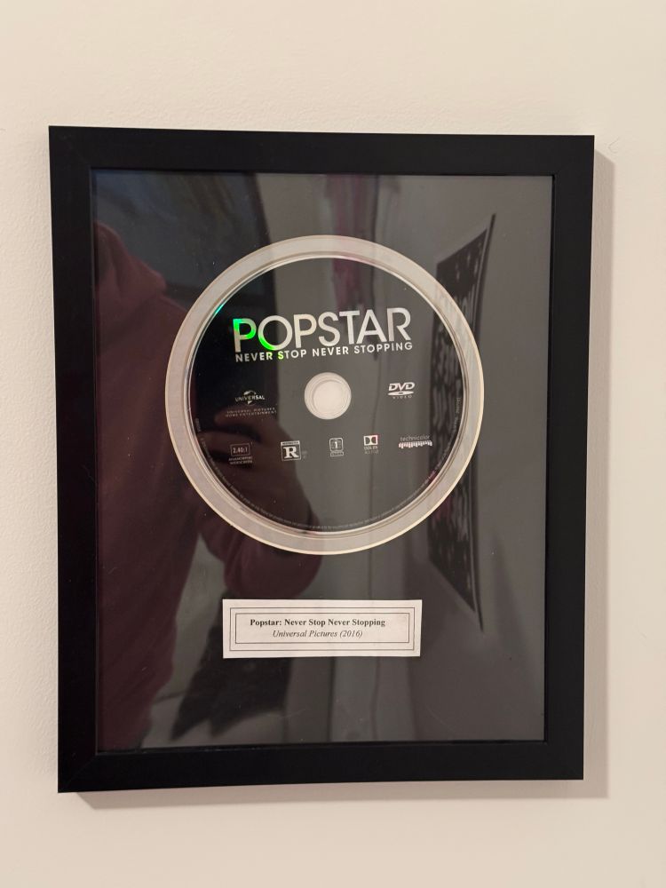 A close up photo of the DVD for Popstar: Never Stop Popping inside a black frame with a label underneath.