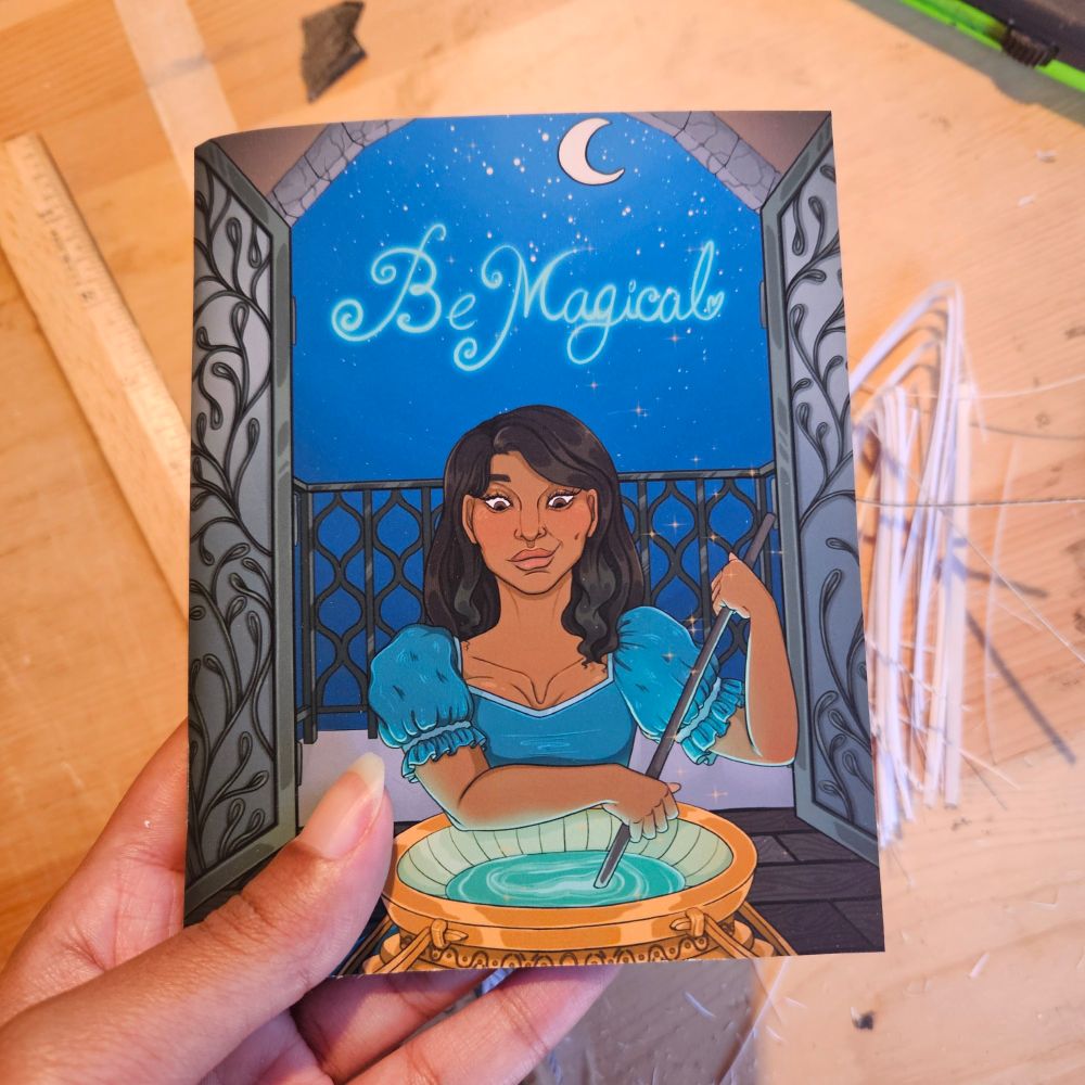 A photo of a pocket journals with my art on it. It has the words "Be Magical" with a woman stirring a golden cauldron.