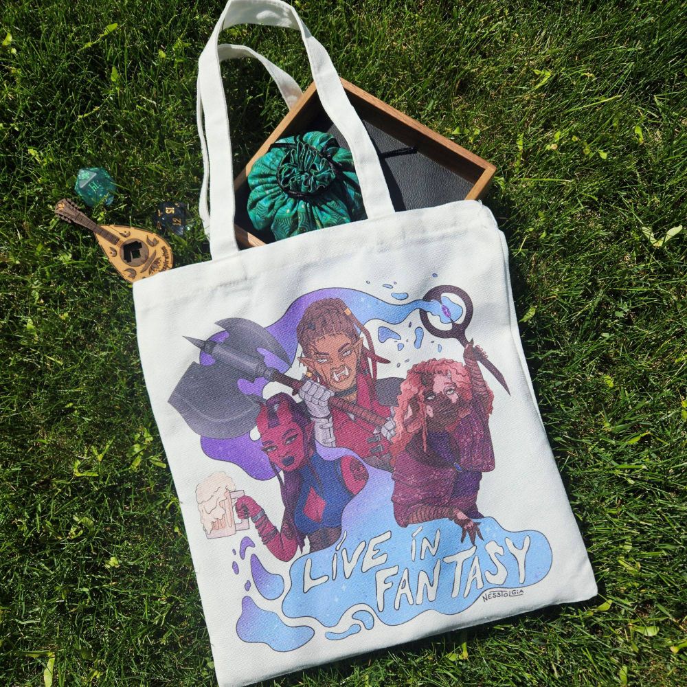 A photo of an tote bag with my art on it. It's of three fantasy characters with the words "Live In Fantasy" underneath. It has a tiefling holding a pint of ale, a half orc holding a great axe, and a firbolg holding up a staff. The magic from the staff swirls around them, which is where the words are written below. 