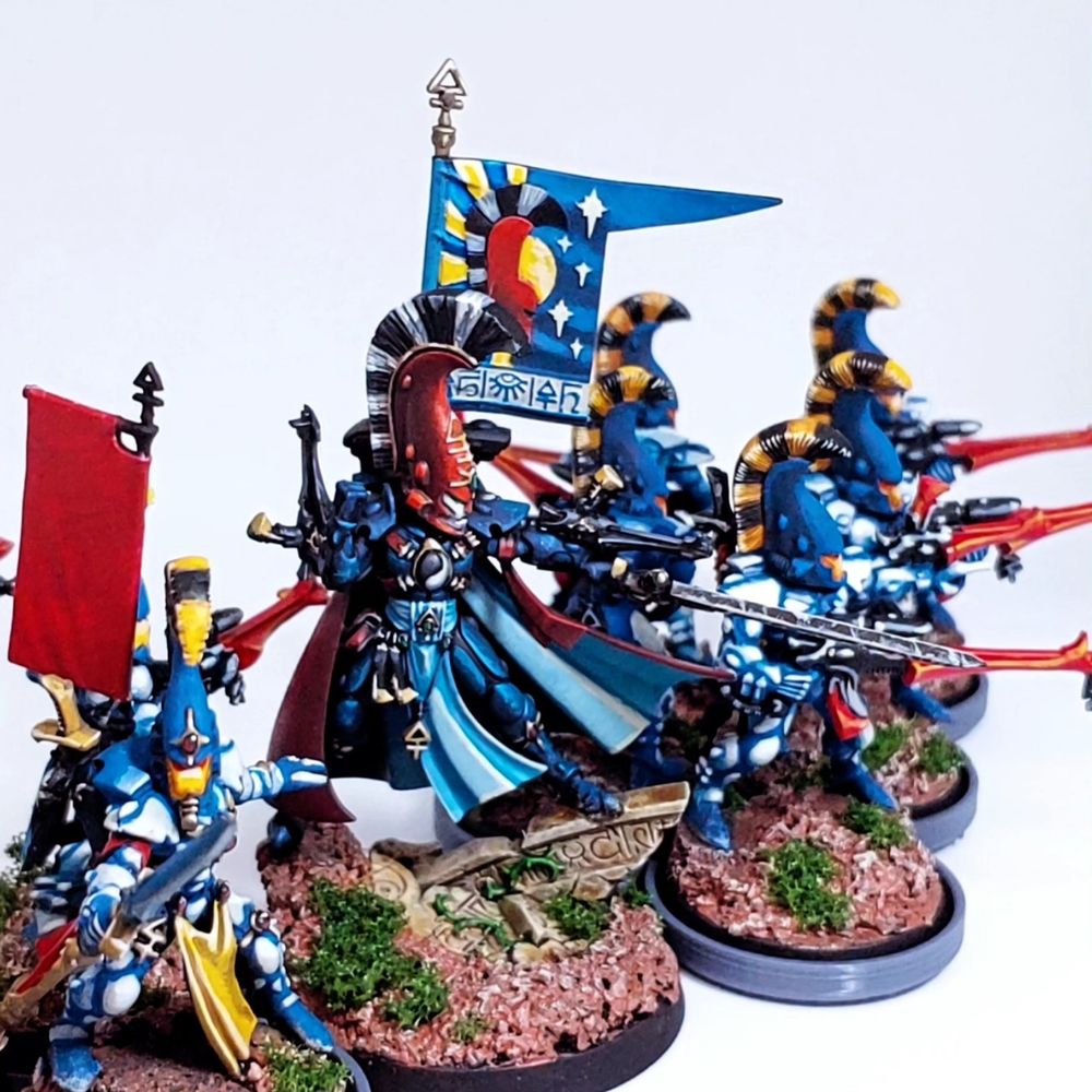 A blue armored eldar with a red helmet leads a squad of similarly colored Eldar riflemen carrying blue shuriken rifles. 