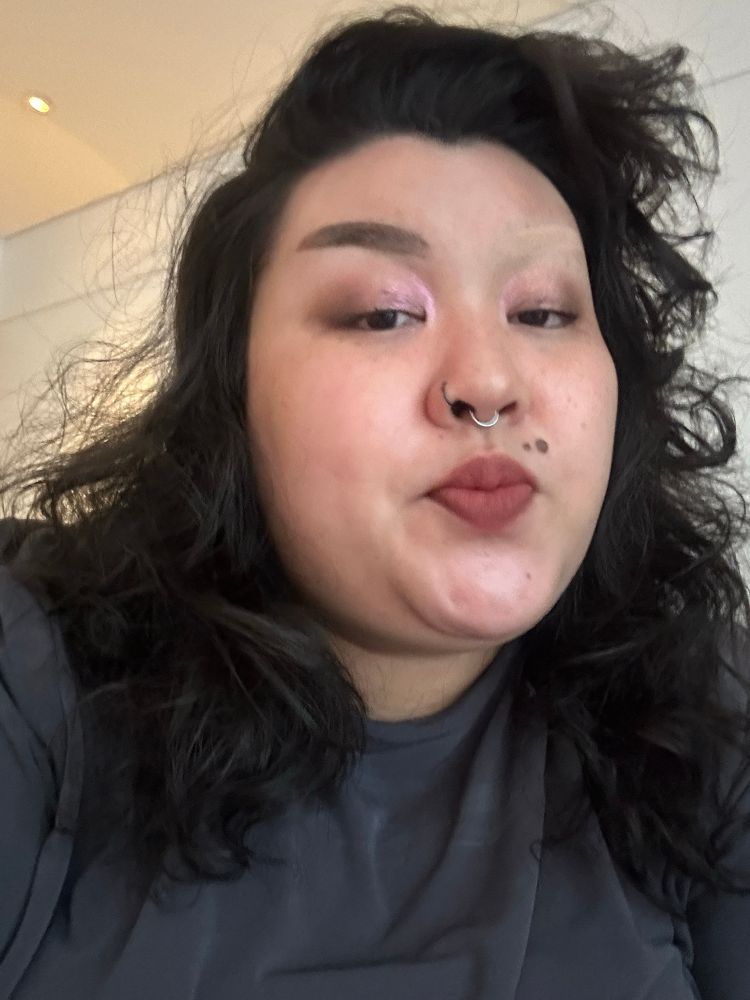 A selfie of Clore. Their hair is down and they have one brunette and one blonde eyebrow. Their mouth is scrunched and they are looking down and off camera. 