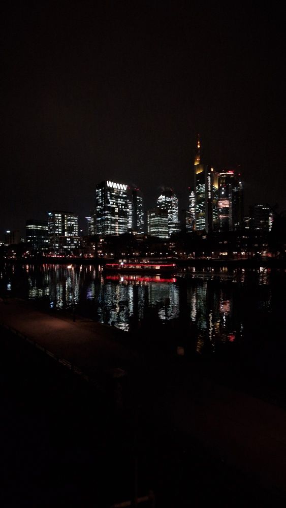 Frankfurt bei Nacht, hell erleuchtete Hochhäuser, reflektiert im Main, über den Fluss fährt ein rot erleuchtetes Schiff, ebenfalls reflektiert.