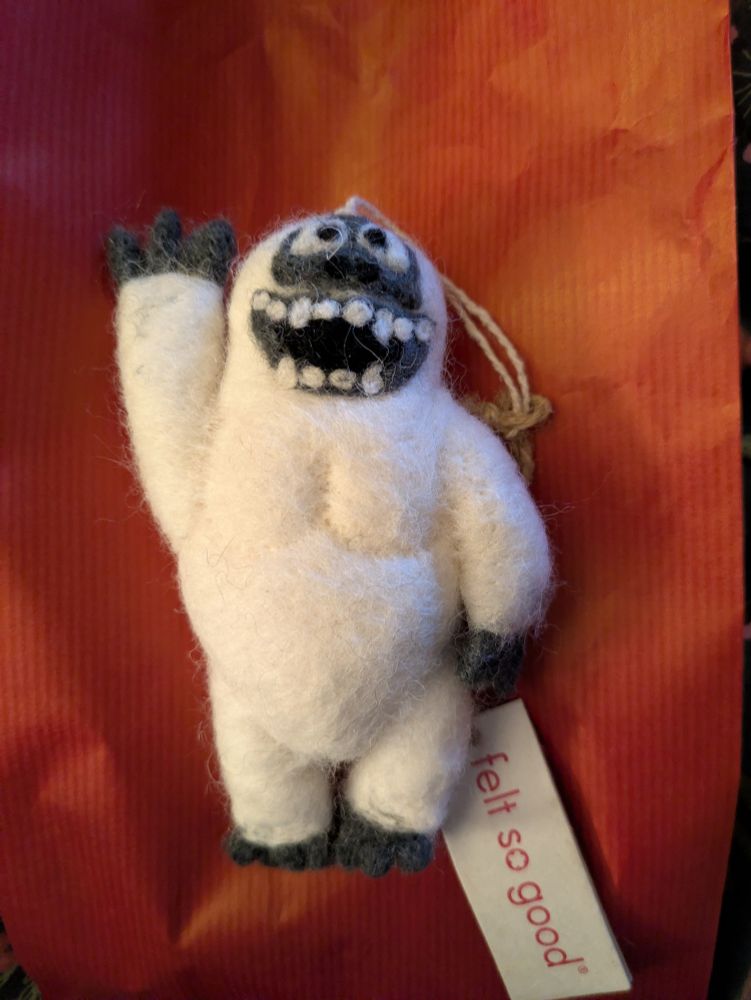A felted yeti. He is mostly white with hands feet and face in grey, with white teeth and eyes. He is smiling and raising his right arm in a wave hello. 
