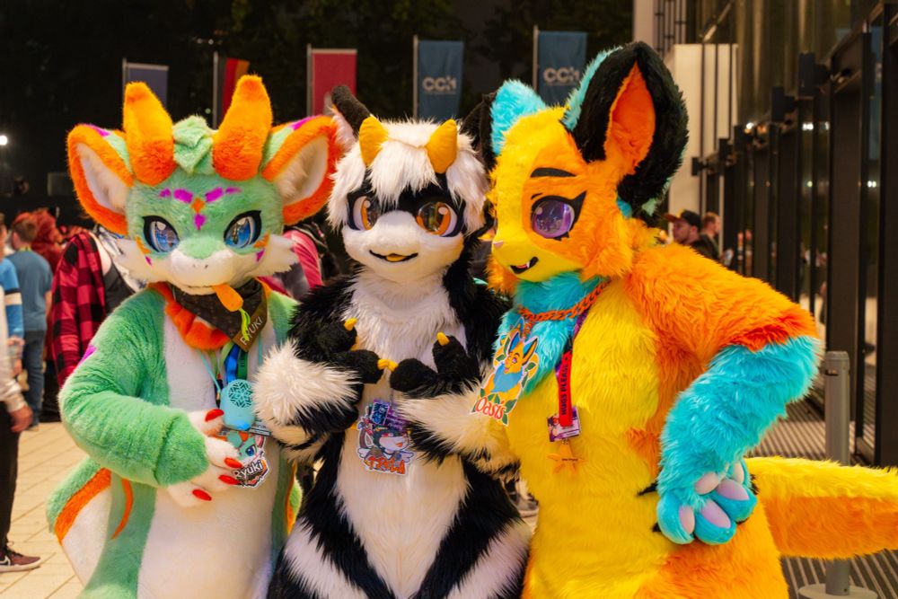 Three colorful fursuiters pose at night during a lively event. On the left is a green and white dragon with orange and purple markings, horns, blue eyes and a carrot in it's mouth. In the middle is a black and white creature with golden eyes. On the right is a vibrant yellow, orange, and turquoise creature with purple eyes. Flags and a crowd of attendees are visible in the background