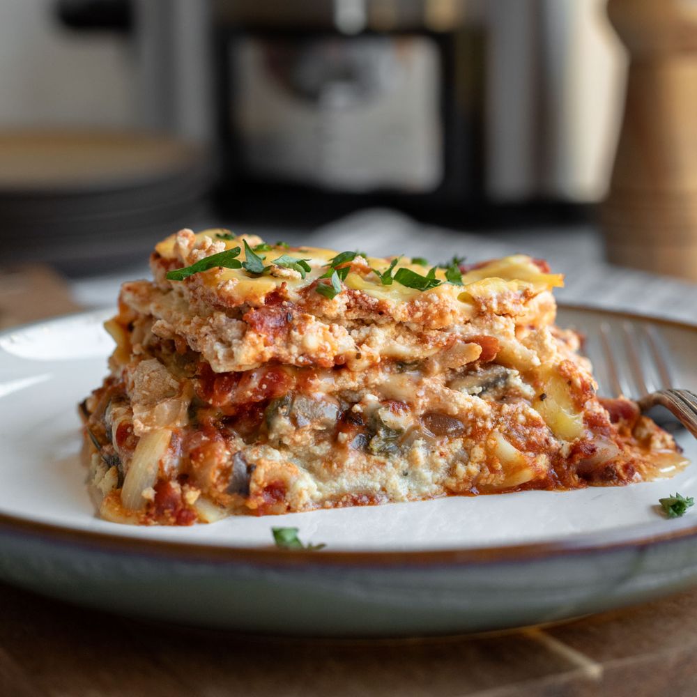 A serving of vegan lasagna on a plate with a slow cooker in the background.