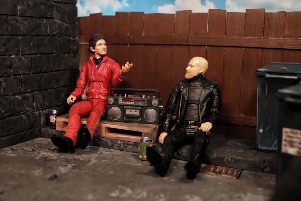 Diorama Two figures sitting on a sidewalk next to a wall, in front of a wooden fence. 