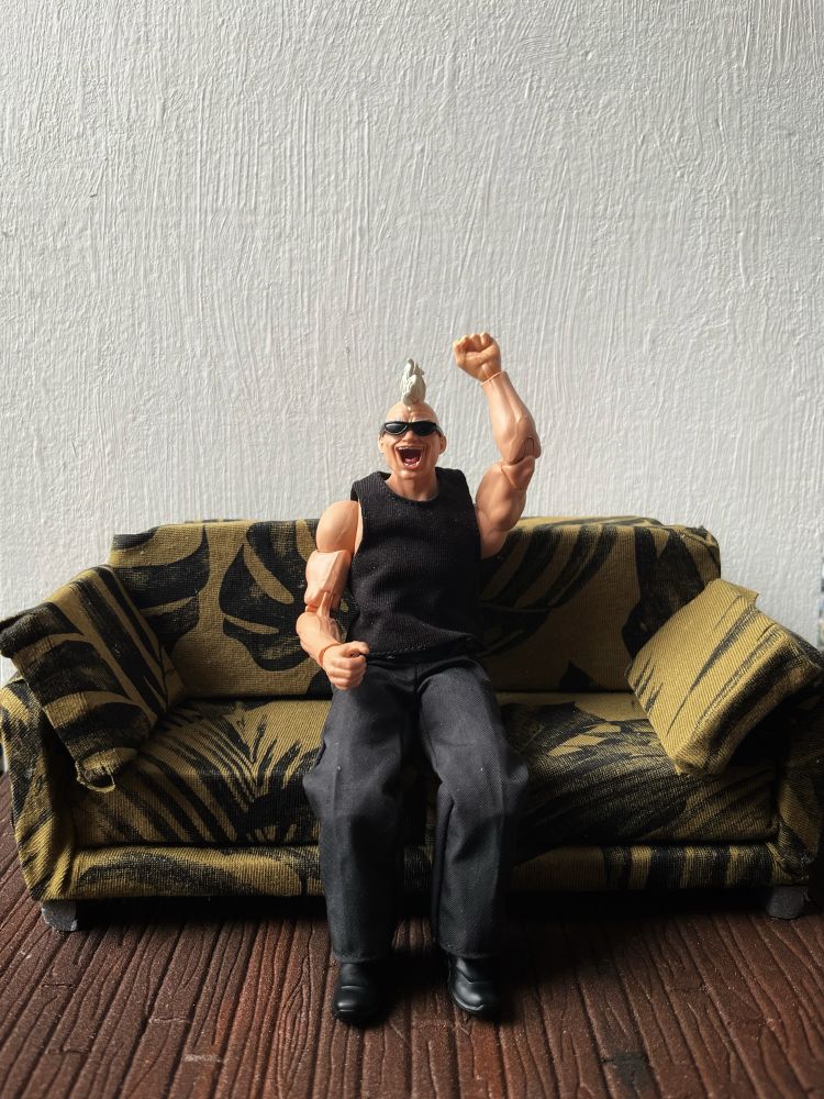 Action figure sitting on a couch that is patterned with green cloth and darker green leaves. The floor is wooden and the background wall is white.