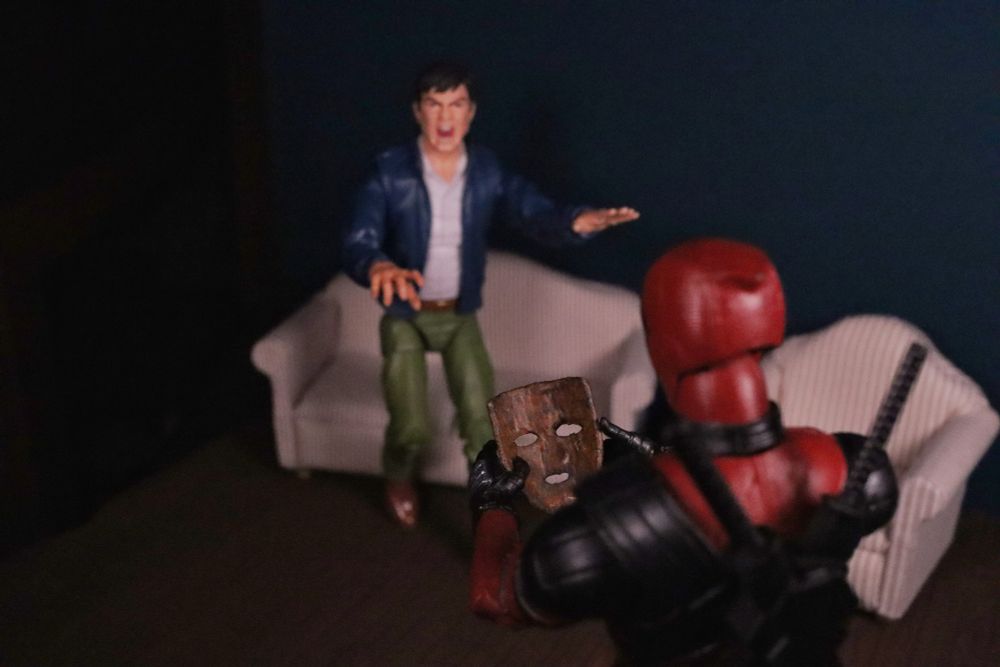 Deadpool holding the wooden mask from the movie The Mask. In the background is a man shouting and holding out his hands in a no, stop gesture 