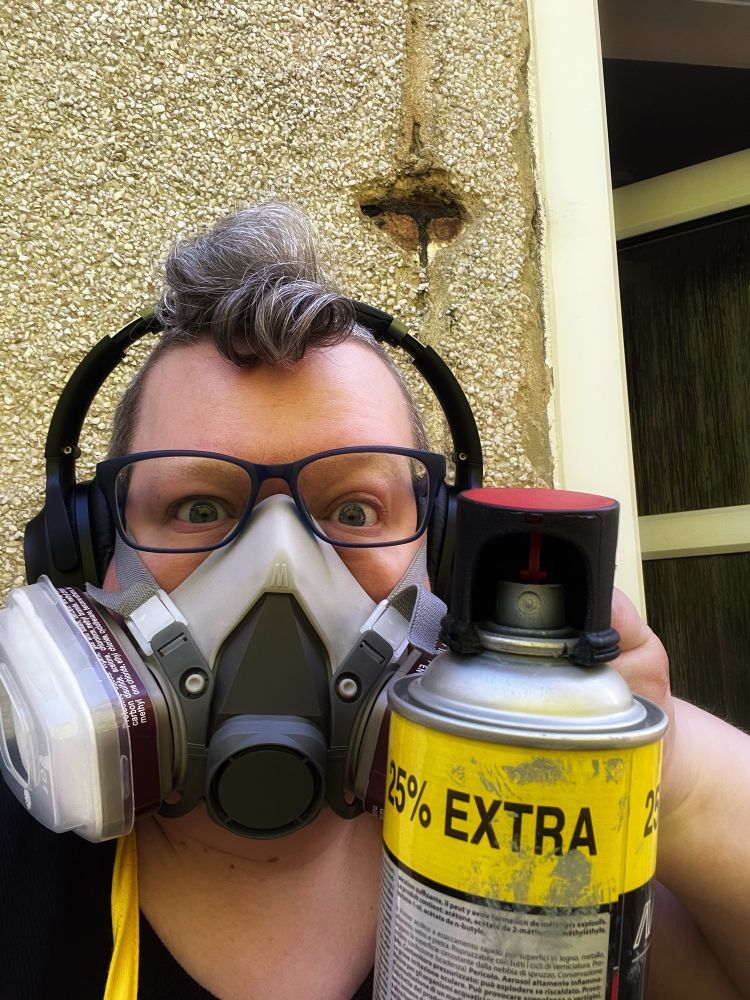 A person with slightly poofy hair on the top. Wearing glasses and a respirator. They're pointing a spray can at the camera. 