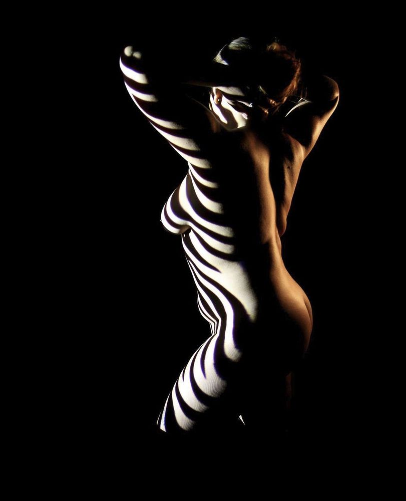 Nude female model, with zebras by bodylighting on the face, chest and leg. Low key light on the back, following the model muscles.
