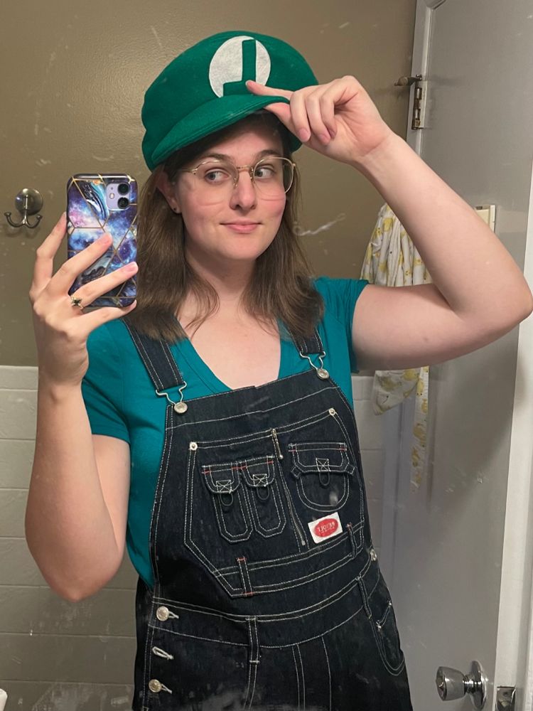 Selfie of me in overalls, a green V-neck T-shirt, and a Luigi hat