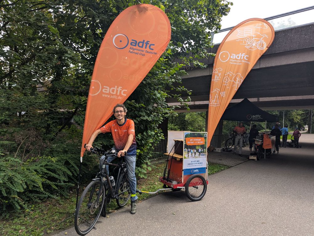 Der ich auf dem Rad mit 🔊🎶 Boombox dran, ne ADFC Dropflag vor mir im Ständer, eine hinter mir am Anhänger.
Am Anhänger die Premiere: A1 Sternfahrt-Plakate im neuen Plakathalter. 🎉
Im Hintergrund Pavillon mit Engagierten beim Codieren unter der Brücke.
