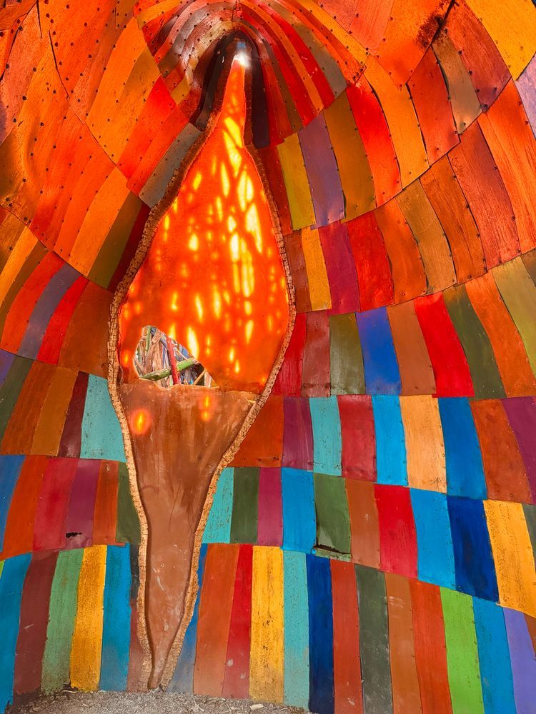 Interior view of a vibrant rainbow-colored wooden structure with curved ribs painted in stripes of red, orange, yellow, blue, and other bright colors, creating a tunnel-like space with light filtering through.