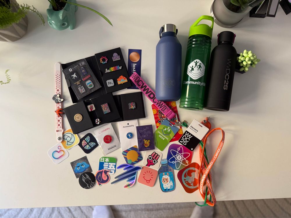 Collection of tech conference swag on a table. Includes water bottles, various pins, badges, stickers with tech logos, and a lanyard. Two small plants are visible in the corners.