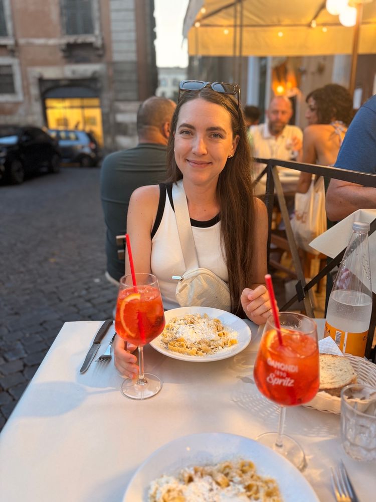 Kathi sitting at an outdoor restaurant table, smiling with a drink and a plate of pasta in front of her.