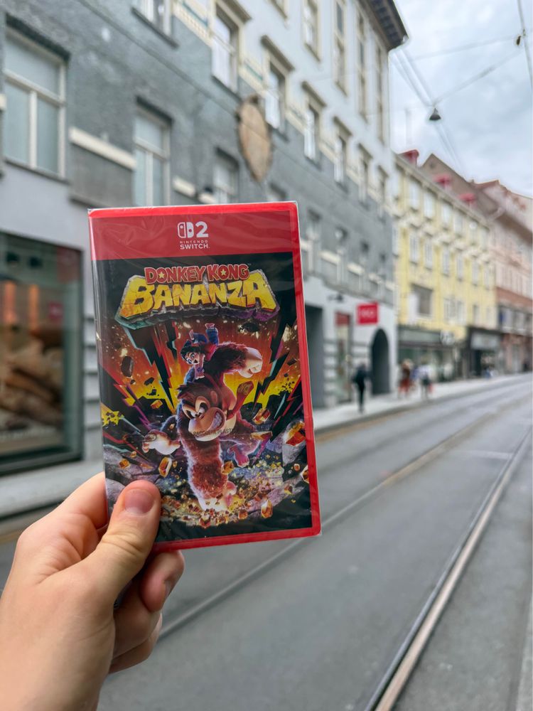Hand holding a Nintendo Switch 2 game titled "Donkey Kong Bananza". Background shows a city street with old buildings, tram tracks, and a cloudy sky.