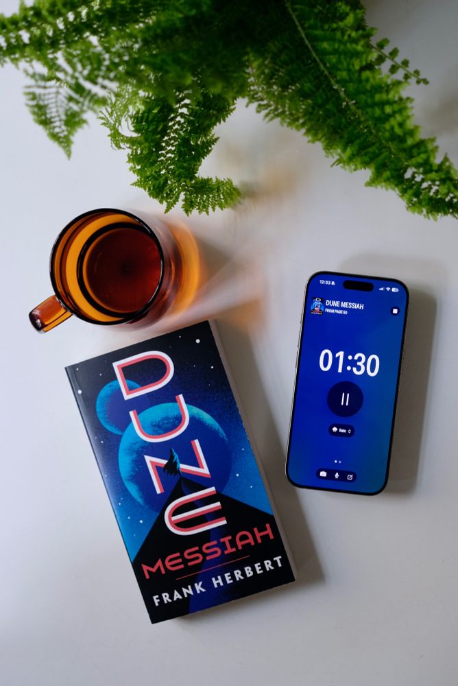 Smartphone running PocketShelf showing a timer counting up. Left of it is a book titled "Dune Messiah" by Frank Herbert on a table next to an amber mug. Green fern leaves hang above.
