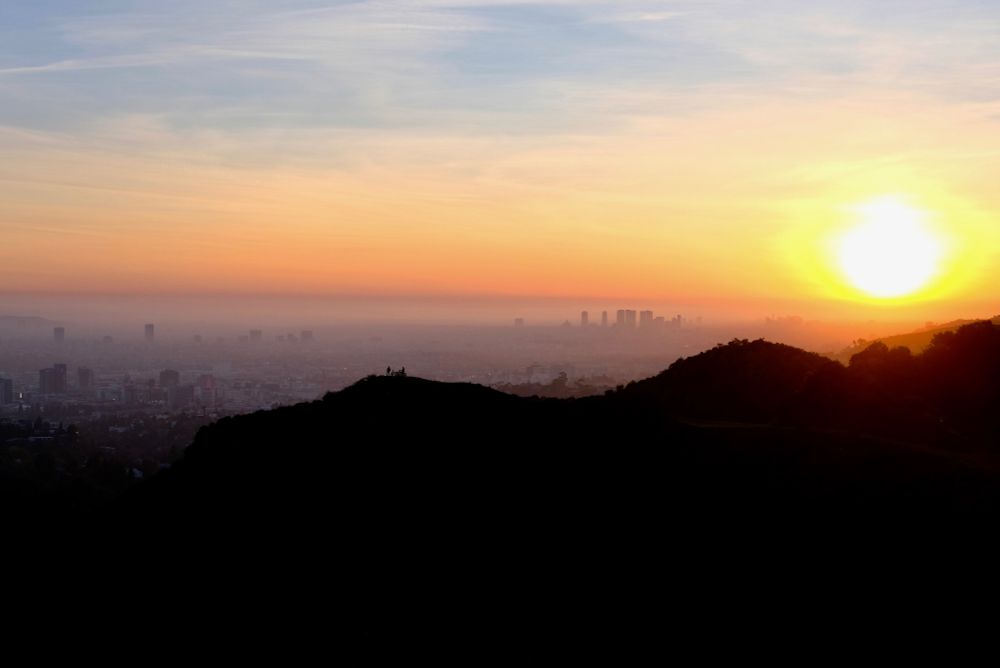 Sunset over LA. You can see buildings in the fog in the background. In the front, there are two hills. On one, two people stand with a bike.