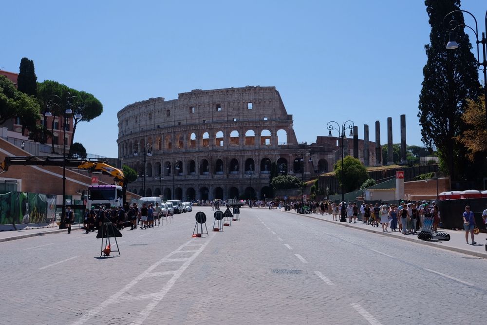 View of the Colosseum in Rome from a street leading up to it. People walk along the sidewalks, and trees and streetlamps line the path.