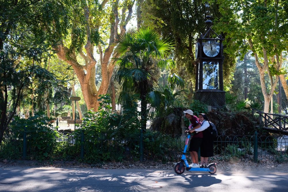 Two people ride a blue electric scooter on a sunny day through a park with lush green trees and a clock tower in the background.