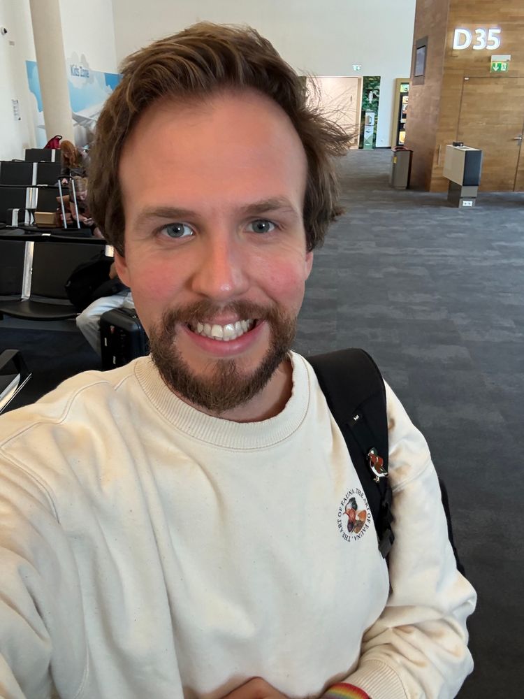 Me smiling at the camera in an airport seating area, wearing a white Art of Fauna sweatshirt and carrying a backpack.