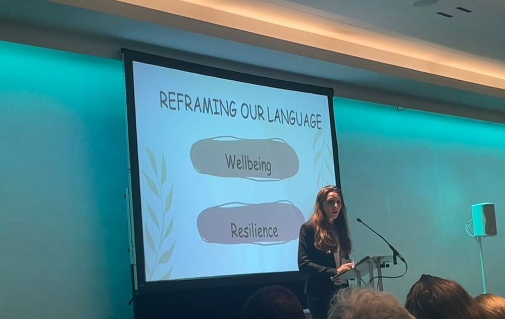 A photo of me speaking at a conference. Behind me is a slide that says reframing our language with the words wellbeing and resilience 