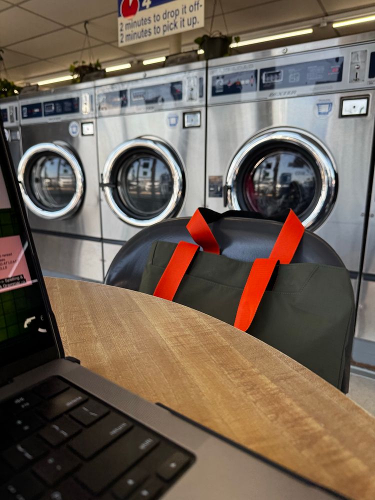 A laptop in the bottom left corner of the screen on a table, a bag in the chair in the center of the frame, and laundry machines in the background 
