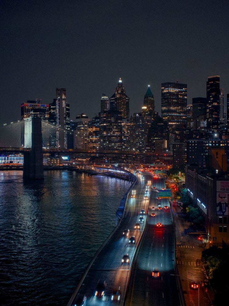 A night view of the Brooklyn Bridge and New York City skyline with illuminated buildings and a busy highway, photographed in Manhattan, New York.