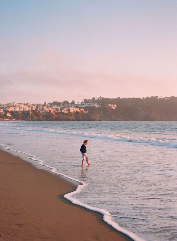 A girl walks along the edge of the ocean as waves gently touch the shore in San Francisco, California. San Francisco, California photographed on medium format film.