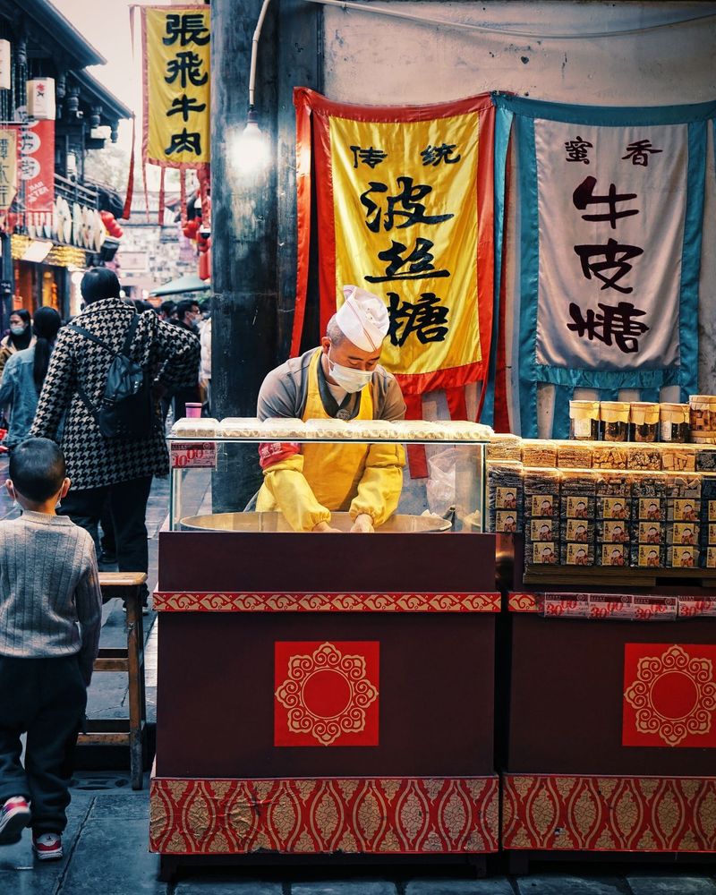 Under a bare bulb that pretends to be a small moon, the candy man kneads winter into sweetness. Banners flutter like stage curtains; the crowd edits itself to background noise. A boy stalls at the edge of the frame. I order something to keep for later, and later slips past—sticky, warm, leaving subtitles on my hands.