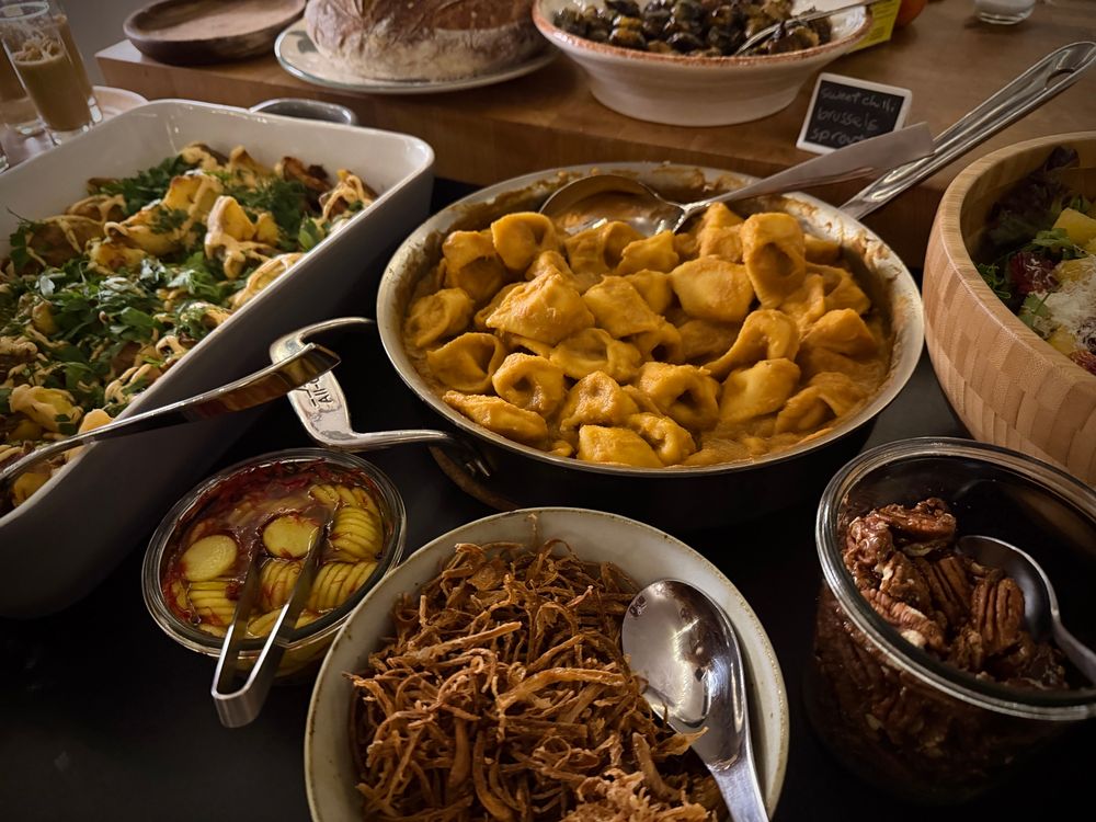 A spread of food in various cookware and serving trays. There are smashed potatoes, butternut squash cheese tortellini, fried shallots, salad and candied walnuts. This is only a portion of what was served to guests.