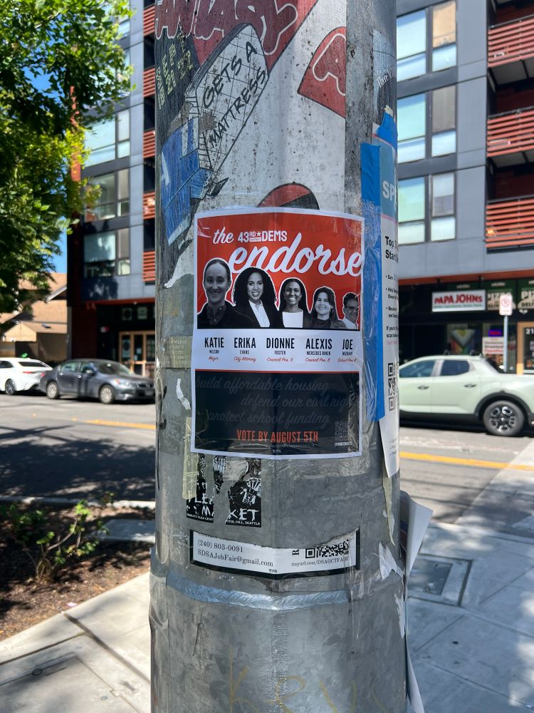 A poster taped to a traffic light pole that lists five candidates endorsed by the 43rd District Democrats: Katie Wilson for Mayor, Erika Evans for City Attorney, Dionne Foster for Council Position 9, Alexis Mercedes Rinck for Council Position 8, and Joe Mizrahi for School Board Position 4.
