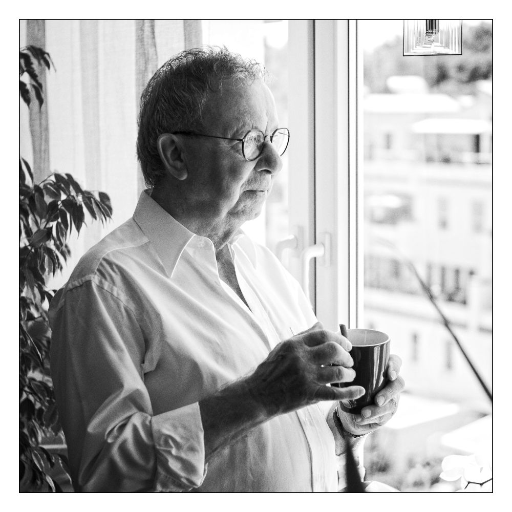 Älterer Herr steht mit einer Kaffeetasse in der Hand innen am Fenster.