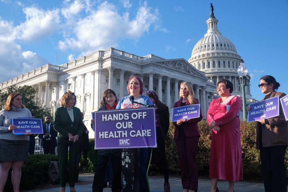 A group of women stand outside the U.S. Capitol holding purple signs reading messages like “Hands off our health care” and “Save our health care.” One woman speaks at a podium as others stand in solidarity around her under a sunny sky.