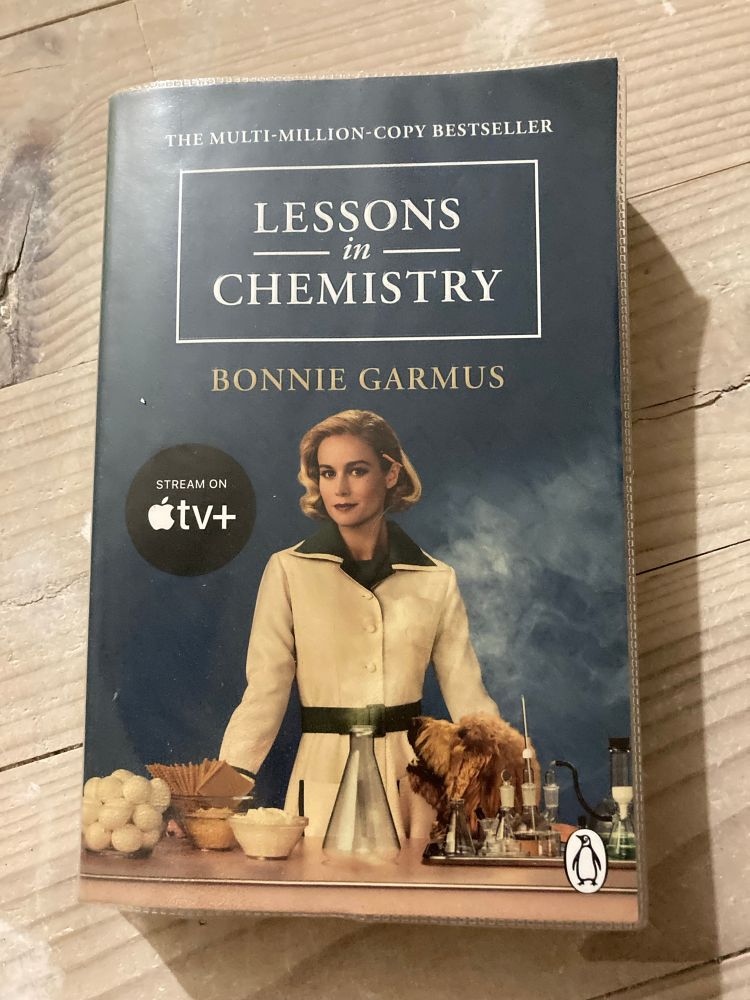 Book cover of Lessons in Chemistry by Bonnie Garmus shoeing a woman in a front of a table with food.
