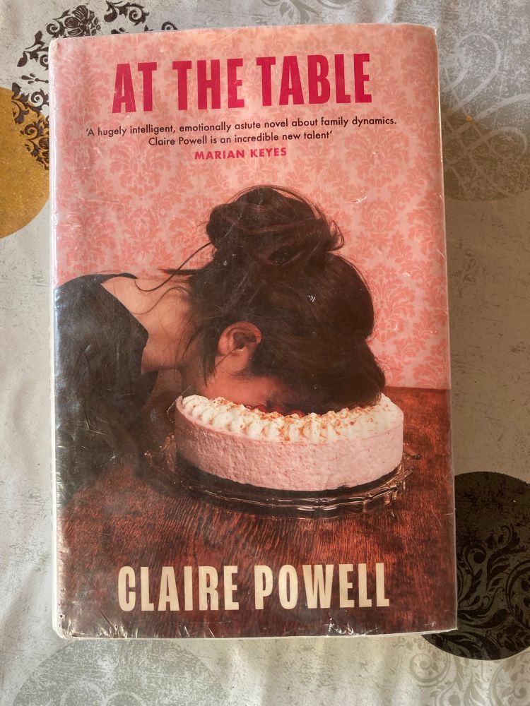 Book cover of At the Table by Claire Powell showing a girl with her face in a cake