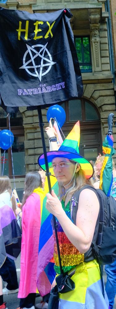 Picture take of me at a Pride Parade. I wear a nonbinary flag as skirt, combined with a rainbow colored sparkly top. I also sport a witch-inspired pointy hat in rainbow colors. I hold a banner that says "Hex the Patriarchy".