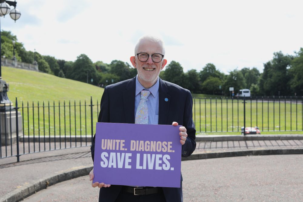 Stewart Dickson MLA, smiling, standing in front of Stormont lawn holding a purple sign reading "Unite. Diagnose. Save lives."