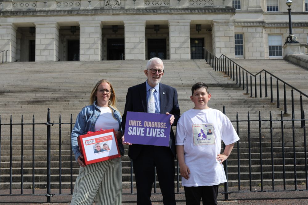Stewart Dickson MLA standing in front of Stormont steps holding a purple sign reading "Unite. Diagnose. Save lives." On his left stands Isla Gear, holding a red box with information about the campaign on the front, and Max on his right.