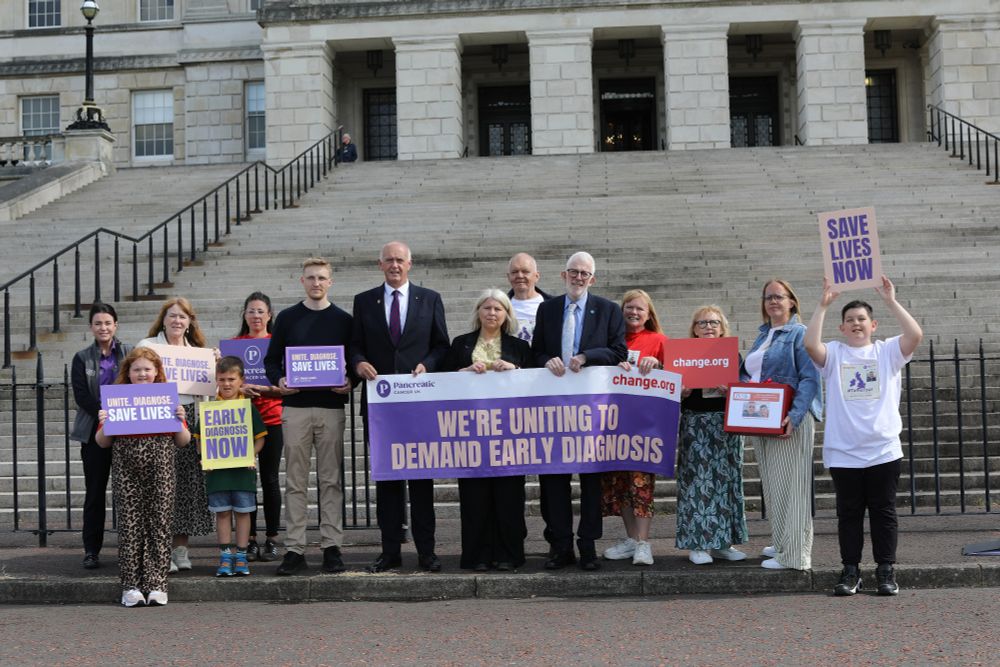 Stewart Dickson MLA standing in front of Stormont steps helping to hold a purple banner reading "We're uniting to demand early diagnosis," surrounded by Isla Gear, and members of both her family and Pancreatic Cancer UK, holding variety of signs.