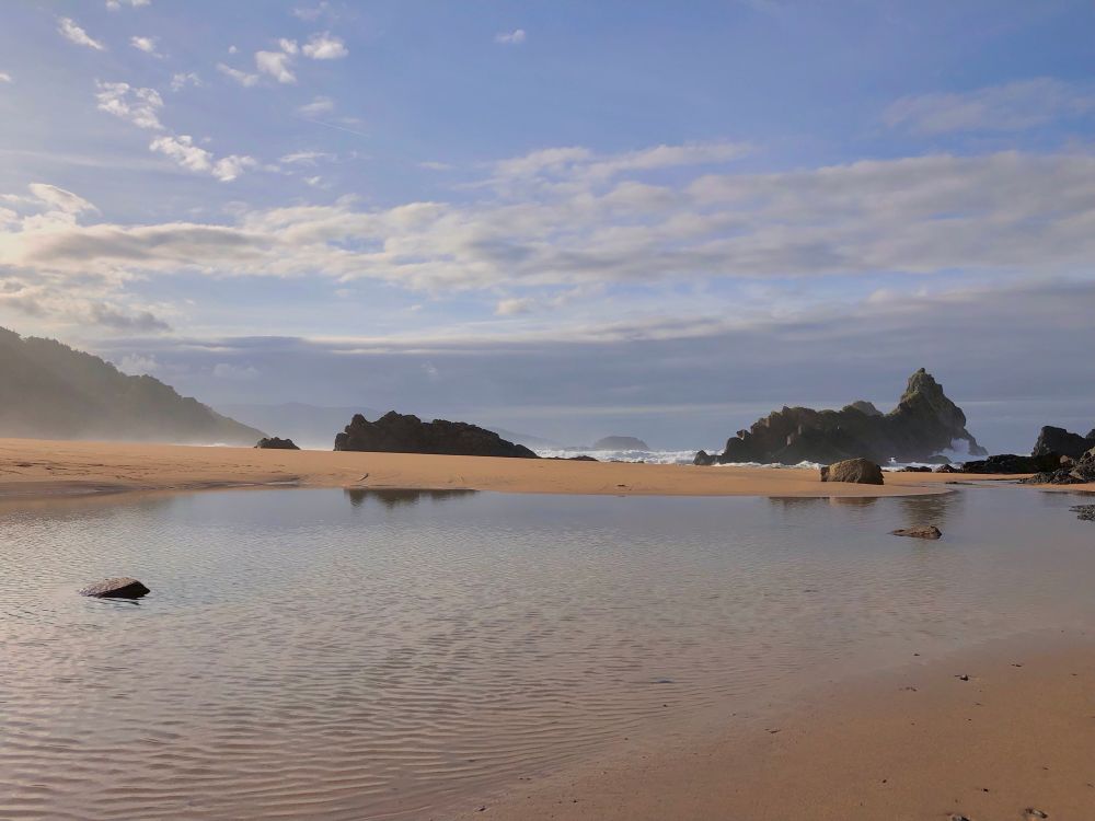 Playa en bajamar, con una lengua de agua entre la arena que refleja las rocas que emergen en el borde de la orilla. El día ha sido lluvioso pero un oportuno claro aporta luz a parte del cielo y a la bruma baja de las olas sobre el perfil de Mundaka. Al fondo se intuye la isla Izaro.