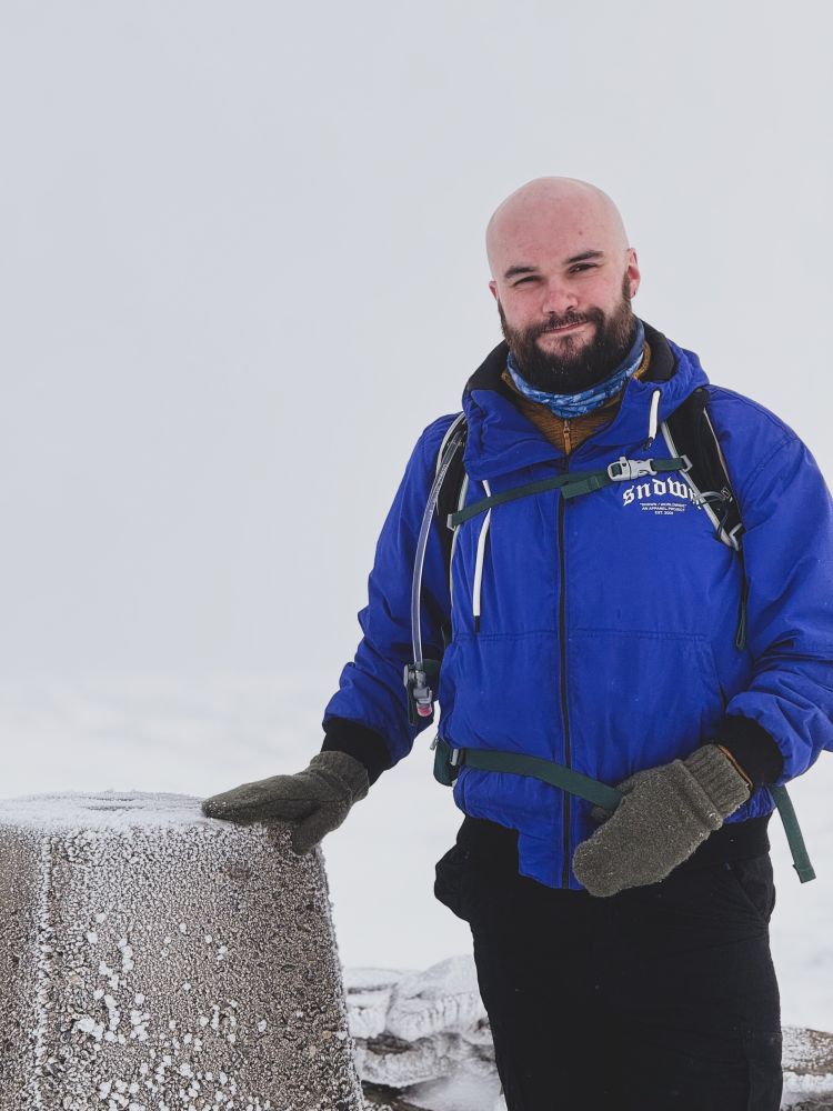 Me standing on a snowy mountain top, my hand on the trig point