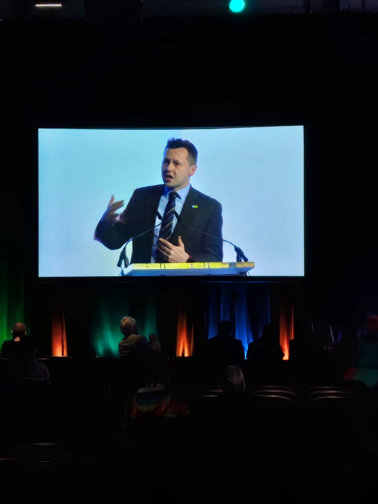 Photograph of a screen showing a white man in a dark blue suit delivering a speech