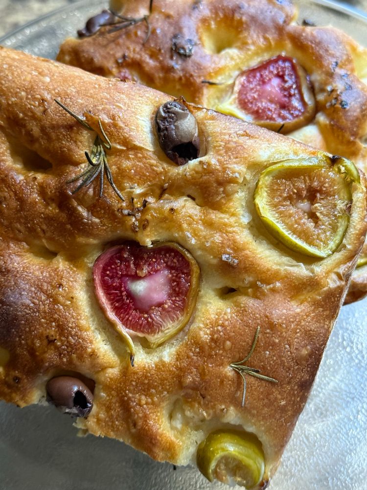 Two squares of home baked focaccia piled on top of each other. The focaccia has little sprigs of rosemary, olives and slices of figs on it. 