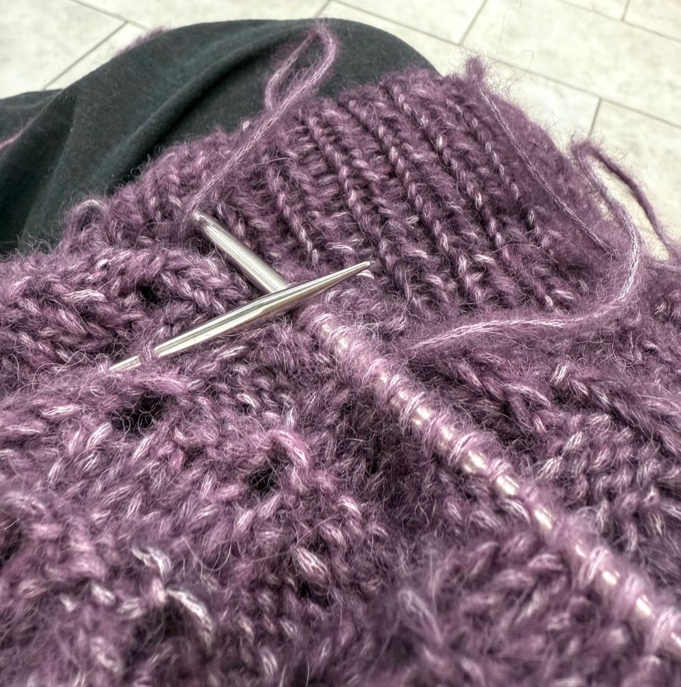 A knitting project is shown in close up. The yarn is a subdued purple and the yarn is a fuzzy yarn with a white cotton core.