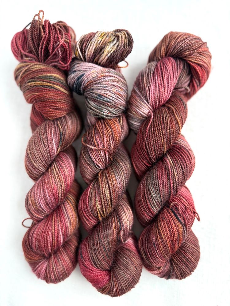 Three skeins of yarn lying on a white background. They are hand dyed in shades of cranberry, gray, brown, gold. 