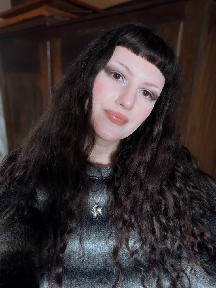 Image is a selfie taken in front of a wooden wardrobe. My hair is long, dark brown, and wavy, and my bangs are quite short. My makeup is rather natural looking, a soft sparkly smokey eye, light peachy brown lip, with blush and highlights. I'm wearing a grey, black, and white striped jumper, and a necklace with multiple small silver pendants visible.   I have a slight friendly smile. 