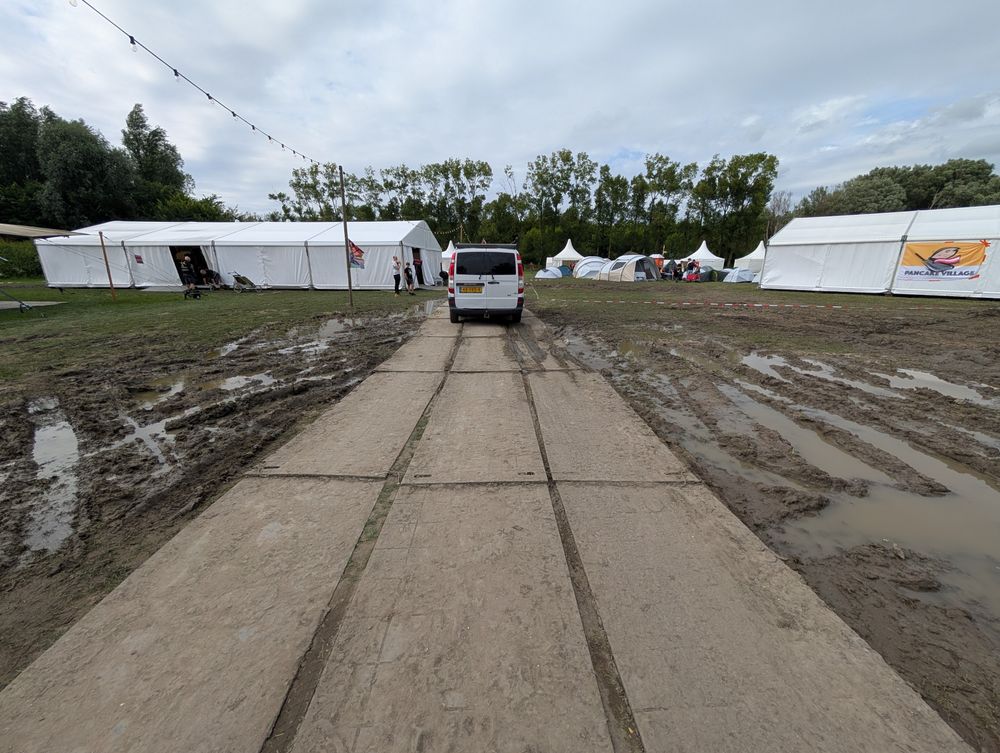 A van drives along road plates (supported by sand) towards a tent. On either side is mud with standing water 