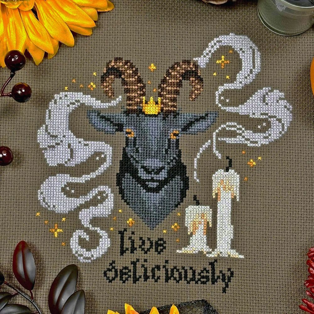 A cross stitch of a black goat face with white candles next to him that have smoke surrounding the goat. Text below him says "Live Deliciously"
Goat wear a small gold crown with gold decorative elements scattered around. 